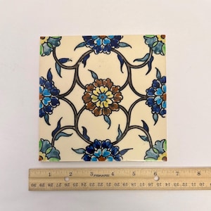 May include: Square ceramic tile with a floral design. The tile features a cream-colored background with a repeating pattern of blue, brown, and yellow flowers connected by dark brown stems. The tile measures approximately 8 inches square.