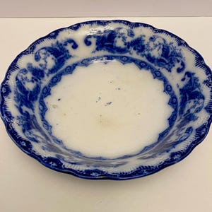 May include: An antique ceramic bowl in white and blue, with a scalloped edge. The rim is decorated with a detailed blue floral and figurative design. The centre of the bowl is white.