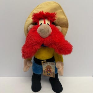 Può includere: Un peluche di Yosemite Sam, un personaggio dei cartoni animati, con cappello giallo, camicia, pantaloni blu e stivali neri. Il giocattolo ha grandi baffi rossi e un'etichetta con la scritta "Yosemite Sam".