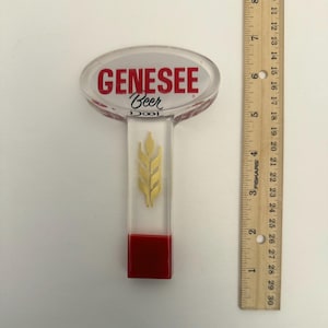 Peut inclure: Un pommeau de robinet de bière transparent avec le texte "GENESEE Beer" en rouge. La poignée a un motif d'épi de blé doré et une base rouge. Une règle est à côté de la poignée, mesurant en centimètres.