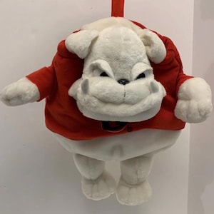 May include: A plush bulldog toy with white fur and a determined facial expression, wearing a bright red short-sleeved shirt. The shirt features a black letter G logo on the chest. The stuffed animal has a round body shape, floppy ears, and visible paws.