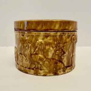 May include: A brown ceramic jar with a lid, featuring embossed jungle-themed designs. The jar's exterior showcases an elephant, palm trees, and other tropical elements. The jar is cylindrical and has a matching lid.