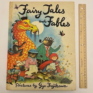 Puede incluir: Una portada de libro infantil vintage con el título "Fairy Tales and Fables" en letras negras. La portada presenta una ilustración colorida de un dragón, un oso, un conejo y otros animales en un entorno de bosque caprichoso. El libro está ilustrado por Gyo Fujikawa.