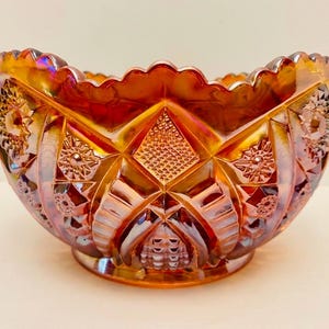 Puede incluir: Un cuenco de vidrio de carnaval iridiscente naranja con un borde festoneado y un patrón prensado de formas de diamante, estrellas y columnas estriadas verticales. El vidrio muestra un brillo metálico reflectante con reflejos púrpuras y dorados.