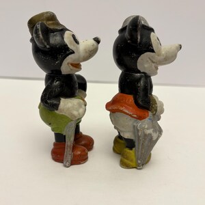 May include: Two vintage Mickey Mouse figurines. One figure wears a hat, green shorts, and red shoes, holding a cane. The other figure wears a hat, orange skirt, and yellow shoes, holding an umbrella. Both figures have black bodies and white faces.