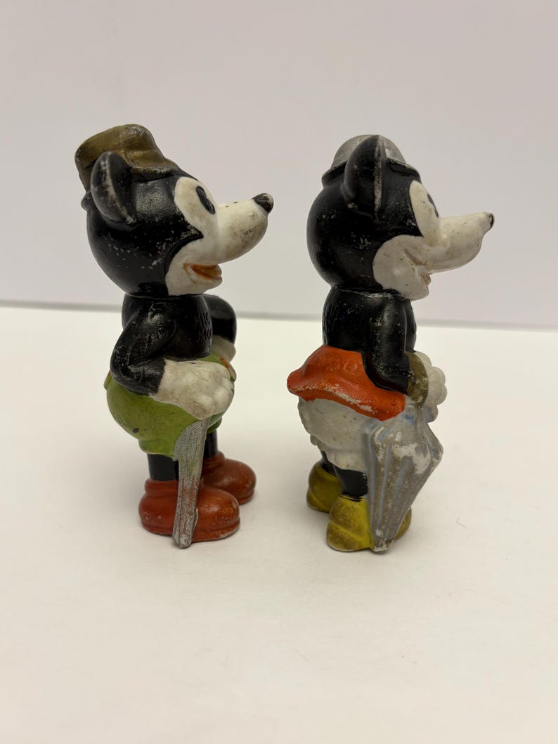 May include: Two vintage Mickey Mouse figurines. One figure wears a hat, green shorts, and red shoes, holding a cane. The other figure wears a hat, orange skirt, and yellow shoes, holding an umbrella. Both figures have black bodies and white faces.