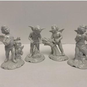 May include: Four white ceramic cherub figurines. Each cherub has wings and is in a different pose, holding various items. The figurines are on a white surface.
