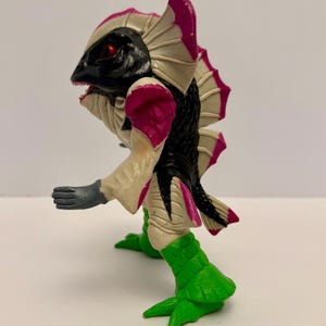 May include: A detailed figurine of a creature with a black head, black and white body, and pink accents. The figure has red eyes, a fish-like head, and green feet. The arms are gray, and the figure is posed as if walking.