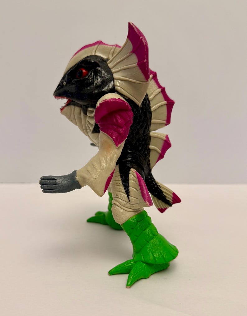 May include: A detailed figurine of a creature with a black head, black and white body, and pink accents. The figure has red eyes, a fish-like head, and green feet. The arms are gray, and the figure is posed as if walking.
