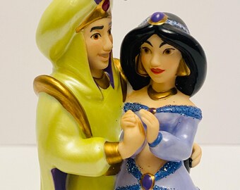 May include: A porcelain figurine of Aladdin and Jasmine from Disney's Aladdin. Aladdin is wearing a yellow robe with a purple turban and Jasmine is wearing a purple dress with blue glitter. They are holding hands and smiling.