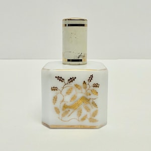 May include: A white, octagonal perfume bottle with a gold floral design and a cylindrical stopper. The stopper is white with gold bands. The bottle's design features gold leaves and a bird-like shape.