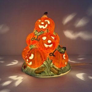 Könnte beinhalten: Eine Keramik-Halloween-Dekoration mit einem Stapel orangefarbener Kürbisse mit geschnitzten Gesichtern, die von innen beleuchtet werden. Die Kürbisse stehen auf einer grünen Basis mit Blättern, und ein Netzkabel ist sichtbar.