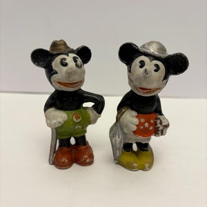 May include: Two vintage Mickey Mouse figurines. One Mickey Mouse figure wears a brown hat, green shorts, red shoes, and holds a silver cane. The other Mickey Mouse figure wears a silver hat, yellow shoes, and holds a red and white polka-dotted object.