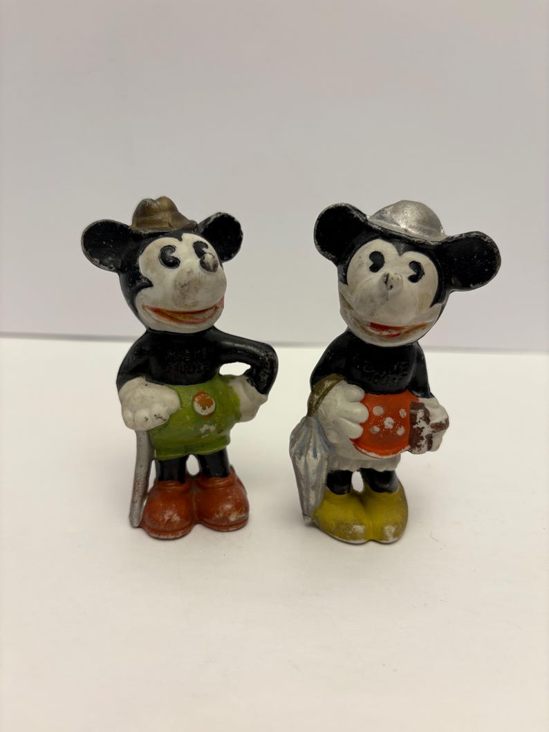 May include: Two vintage Mickey Mouse figurines. One Mickey Mouse figure wears a brown hat, green shorts, red shoes, and holds a silver cane. The other Mickey Mouse figure wears a silver hat, yellow shoes, and holds a red and white polka-dotted object.
