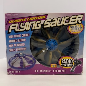 May include: A boxed remote control Flying Saucer toy. The box features the text "Remote Control Flying Saucer" in large letters. The toy has a blue and gold color scheme and includes a remote control. The box also states "Radio Remote Control!" and "No Assembly Required!"