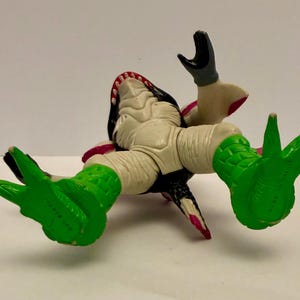 May include: A toy figure of a creature with a white body, black head, and green feet. The figure has an open mouth with visible teeth and is lying on its back. The figure's arms are raised, and the feet are splayed.