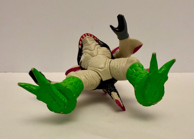 May include: A toy figure of a creature with a white body, black head, and green feet. The figure has an open mouth with visible teeth and is lying on its back. The figure's arms are raised, and the feet are splayed.