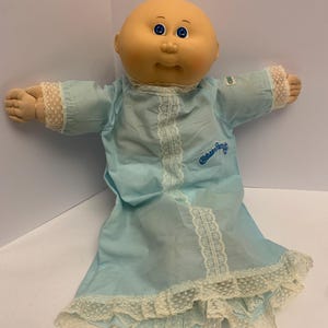 Pode incluir: Uma boneca Cabbage Patch Kid careca vestindo um vestido azul claro com acabamento de renda branca. A boneca tem olhos azuis e uma tez rosada. O vestido tem as palavras "Cabbage Patch Kids" bordadas na frente.