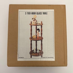 May include: A cardboard box featuring a printed image of a three-tiered hour glass table. The table is brown with decorative spindles and holds small figurines and vases. The text "3 TIER HOUR GLASS TABLE" is printed at the top.