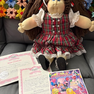 May include: A Cabbage Patch Kids doll with brown yarn hair, blue eyes, and a red, green, and white plaid dress with white lace trim. The doll is accompanied by a birth certificate, official adoption papers, and a colorful birthday card.
