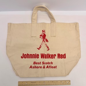 K&ouml;nnte beinhalten: Cremefarbene Canvas-Tragetasche mit rotem Text und einer roten Illustration eines gehenden Mannes. Der Text lautet "Johnnie Walker Red, Best Scotch Ashore & Afloat". Die Tasche hat zwei Griffe.