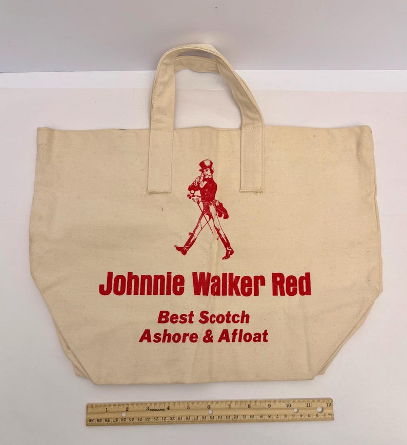 K&ouml;nnte beinhalten: Cremefarbene Canvas-Tragetasche mit rotem Text und einer roten Illustration eines gehenden Mannes. Der Text lautet "Johnnie Walker Red, Best Scotch Ashore & Afloat". Die Tasche hat zwei Griffe.