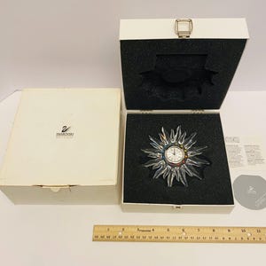 Puede incluir: Una caja blanca con el logotipo de Swarovski y un reloj de cristal en forma de sol con una esfera de colores. El reloj está en una caja blanca con un interior negro.