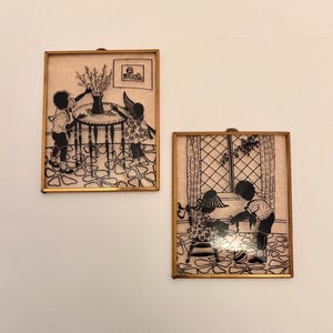 Può includere: Due opere d'arte a silhouette incorniciate. Una mostra due bambini attorno a un tavolo con un vaso di fiori. L'altra mostra due bambini che giocano con una palla vicino a una finestra. Entrambe sono in cornici dorate.