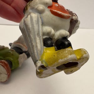 May include: Two vintage ceramic figurines. One figurine is a duck with a white body, orange beak, and black shoes. The other figurine is a green and red figure. Both figurines are on a white surface.