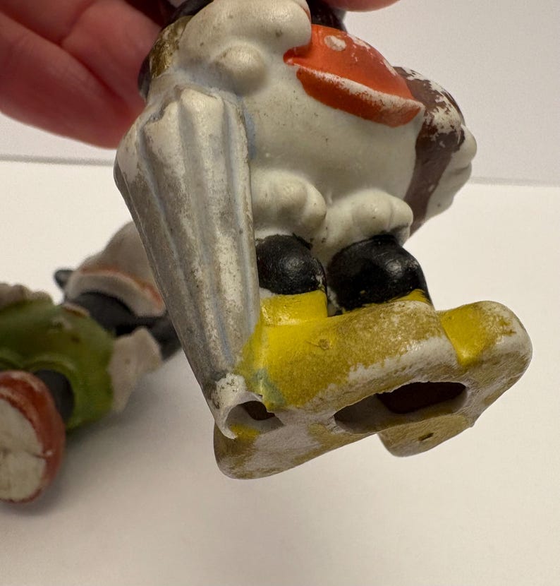 May include: Two vintage ceramic figurines. One figurine is a duck with a white body, orange beak, and black shoes. The other figurine is a green and red figure. Both figurines are on a white surface.