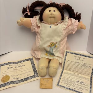 May include: A vintage Cabbage Patch Kid doll with brown yarn pigtails, wearing a pink dress with a white apron featuring a duck design. Includes birth certificate, adoption papers, and name tag.