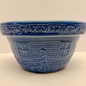 May include: A blue ceramic bowl with an embossed design. The bowl depicts a rural scene with a person watering plants, windows, and floral patterns. The rim has a decorative floral pattern.