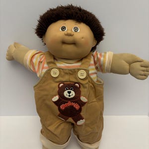 Puede incluir: Una taza de cerámica blanca con asa negra y el texto "But First, Coffee" en negrita negra. La taza se encuentra sobre un fondo borroso, sugiriendo un ambiente acogedor.