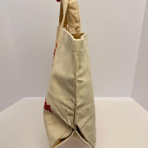 K&ouml;nnte beinhalten: Eine beigefarbene Canvas-Tragetasche mit roter Schrift. Die Tasche hat eine rechteckige Form mit flachem Boden und Henkel. Die Tasche wird von einer Hand gehalten.