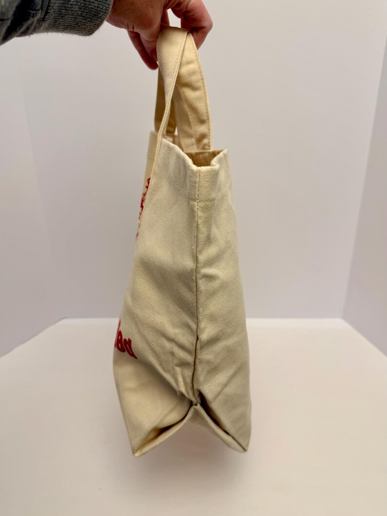 K&ouml;nnte beinhalten: Eine beigefarbene Canvas-Tragetasche mit roter Schrift. Die Tasche hat eine rechteckige Form mit flachem Boden und Henkel. Die Tasche wird von einer Hand gehalten.