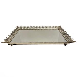 May include: Rectangular vanity tray with a mirrored surface and ornate, filigree-style metal trim. The tray has a cream-colored base and is supported by small feet. The decorative trim features a repeating floral pattern along the edges.