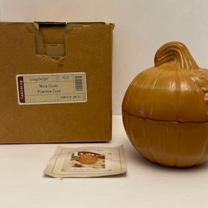 May include: A milk glass pumpkin dish with a lid, in a warm orange hue, sits next to its original cardboard box. The box has a label that reads "Longaberger Milk Glass Pumpkin Dish". A small card with a picture of the dish is also present.