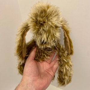 May include: A handmade plush bunny with long, floppy ears and a shaggy, textured fur in shades of brown and tan. The bunny's ears have a contrasting tan fabric lining. The toy is held against a plain white background.