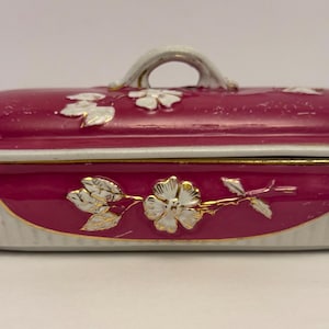 May include: A maroon ceramic trinket box with a lid and handle. The box is decorated with white and gold floral designs. The base of the box is white with gold trim. The handle is white.
