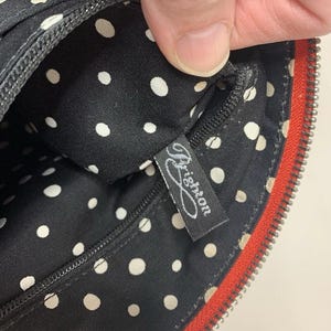 May include: Black handbag interior with white polka dots. The interior features a black zipper and a tag with the word "Brighton" in cursive. The bag has a red zipper trim.