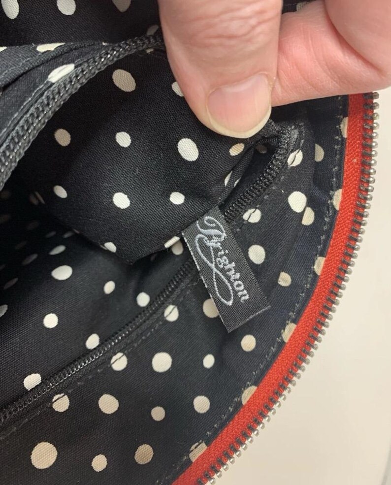May include: Black handbag interior with white polka dots. The interior features a black zipper and a tag with the word "Brighton" in cursive. The bag has a red zipper trim.