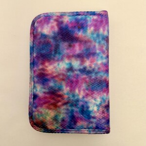 May include: A rectangular case with a rounded top edge, featuring a vibrant tie-dye pattern in shades of purple, blue, and pink. The case has a textured surface and a stitched border, suggesting it's a protective cover for a device or accessory.
