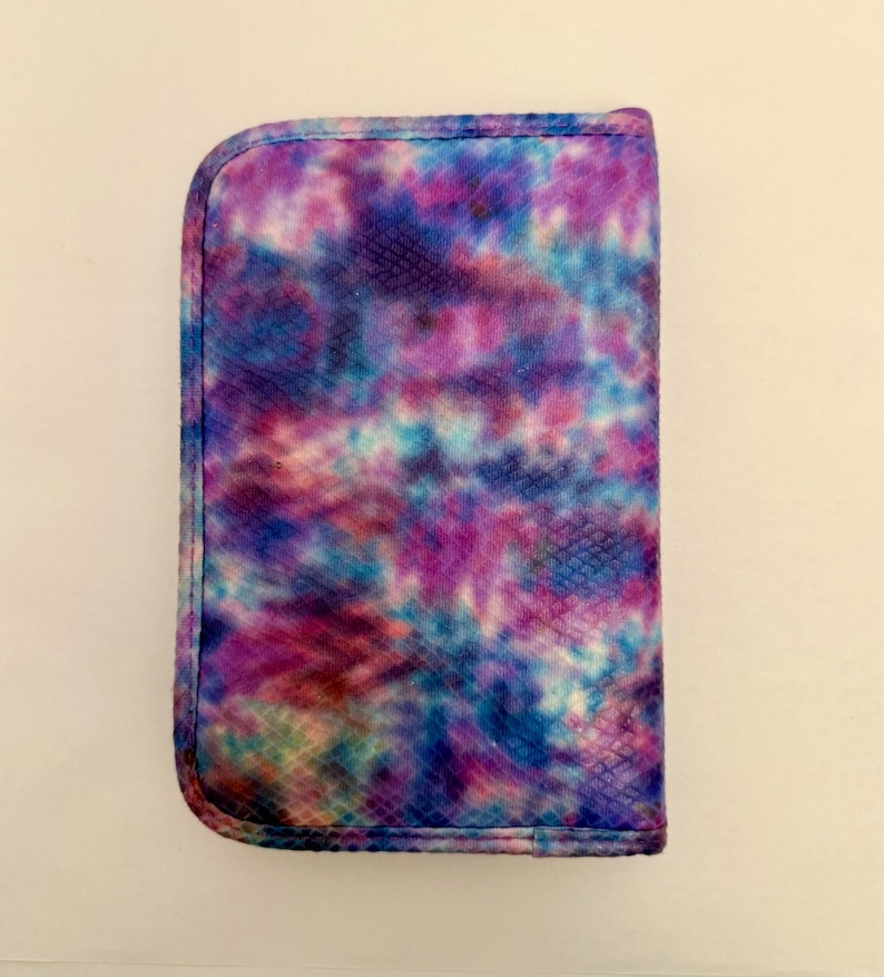 May include: A rectangular case with a rounded top edge, featuring a vibrant tie-dye pattern in shades of purple, blue, and pink. The case has a textured surface and a stitched border, suggesting it's a protective cover for a device or accessory.