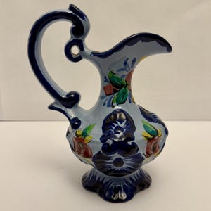 May include: A ceramic pitcher with a light blue body and a dark blue handle and base. The pitcher is decorated with floral patterns in red, green, and yellow. The handle has a decorative, curved design.