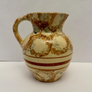 Puede incluir: Un pequeño jarro de cerámica con asa. El jarro tiene una base color crema con patrones abstractos marrones y beige. Dos rayas horizontales marrones rodean la parte inferior. Probablemente se utiliza para servir bebidas.