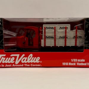 May include: A red and white 1918 Mack flatbed truck model, 1/33 scale, in a display box. The box features the True Value logo and the text "Help Is Just Around The Corner." The model is labeled as an adult collectible.