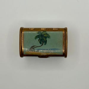 May include: A small, rectangular, vintage pillbox with a hinged lid. The box has a gold-toned metal frame and features a painted scene of a palm tree on a beach with blue water and a light blue sky. The box is closed.