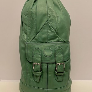 Puede incluir: Una mochila de cuero verde con la parte superior redondeada y un bolsillo delantero con solapa. La mochila tiene hebillas plateadas y un asa en la parte superior. La bolsa mide aproximadamente 51 cm de alto.