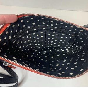 May include: Open view of a small, orange crossbody bag with a black strap. The interior is black with white polka dots and has multiple pockets. The zipper is silver.