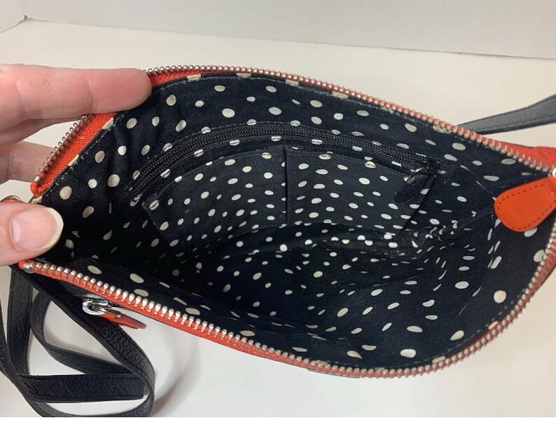 May include: Open view of a small, orange crossbody bag with a black strap. The interior is black with white polka dots and has multiple pockets. The zipper is silver.